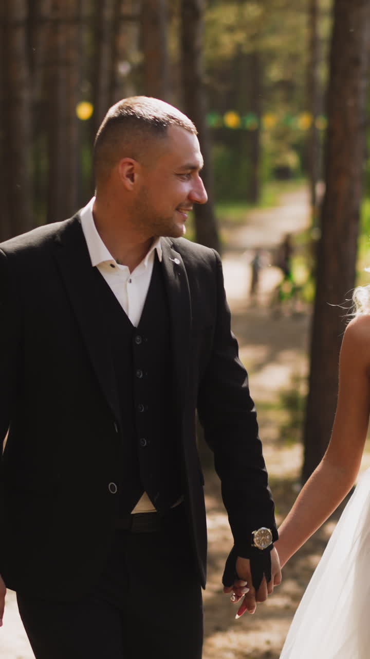 Lovely newlywed couple of happy groom in suit and bride in chiffon wedding dress walks joining hands along green sunny park slow motion
