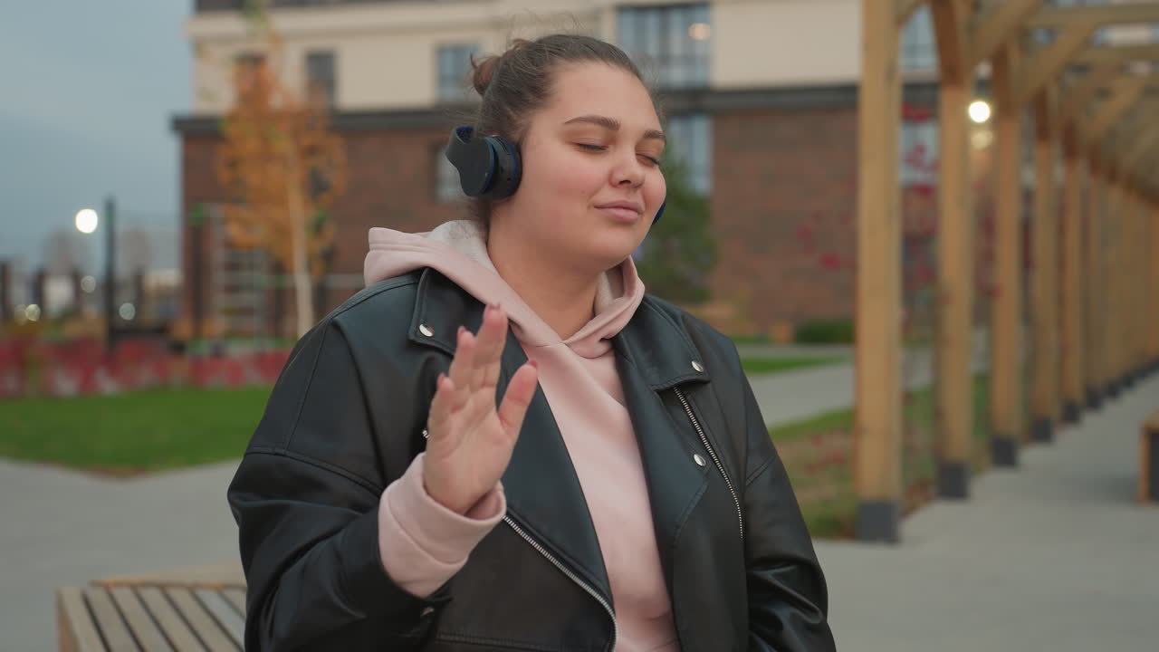 Joyful woman wearing pink hoodie and black leather jacket dances outdoors in urban setting while enjoying music through headphones as distant bokeh lights blink softly behind
