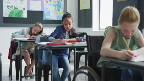 Video-of-caucasian-schoolgirl-in-wheelchair-at-desk-working-in-diverse-in-class,-copy-space