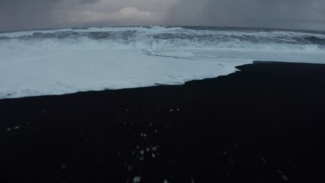 Eine-Wunderschöne-Drohnenaufnahme-Des-Schwarzen-Strandes-In-Island-Zeigt-Die-Hohen-Wellen-Des-Meeres-Und-Das-Bewölkte-Wetter,-Während-Der-Schnee-Einen-Teil-Des-Schwarzen-Sandes-Bedeckt,-Während-Menschen-Stehen