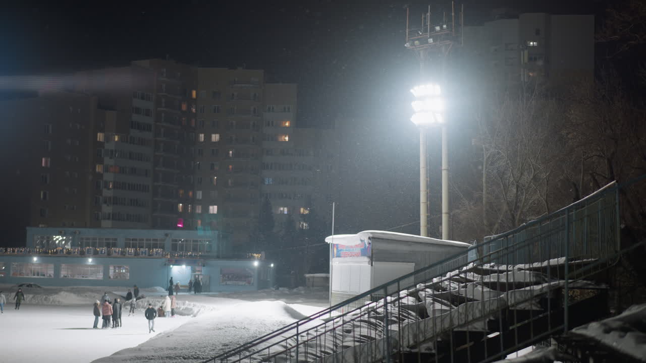 Group of people enjoying winter skating activity on frozen stadium rink at night under bright floodlights, surrounded by snowy environment and tall buildings, with visible falling snowflakes