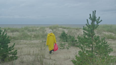 frau in gelbem regenmantel mit roter tasche geht entlang der sanddünen in richtung meer, glatte tracking-schuss