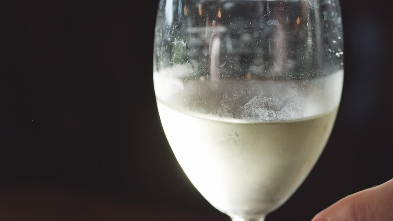 A hand gently lifts a cold white wine glass from a table in a dimly lit, elegant setting. Side lighting highlights condensation and clarity