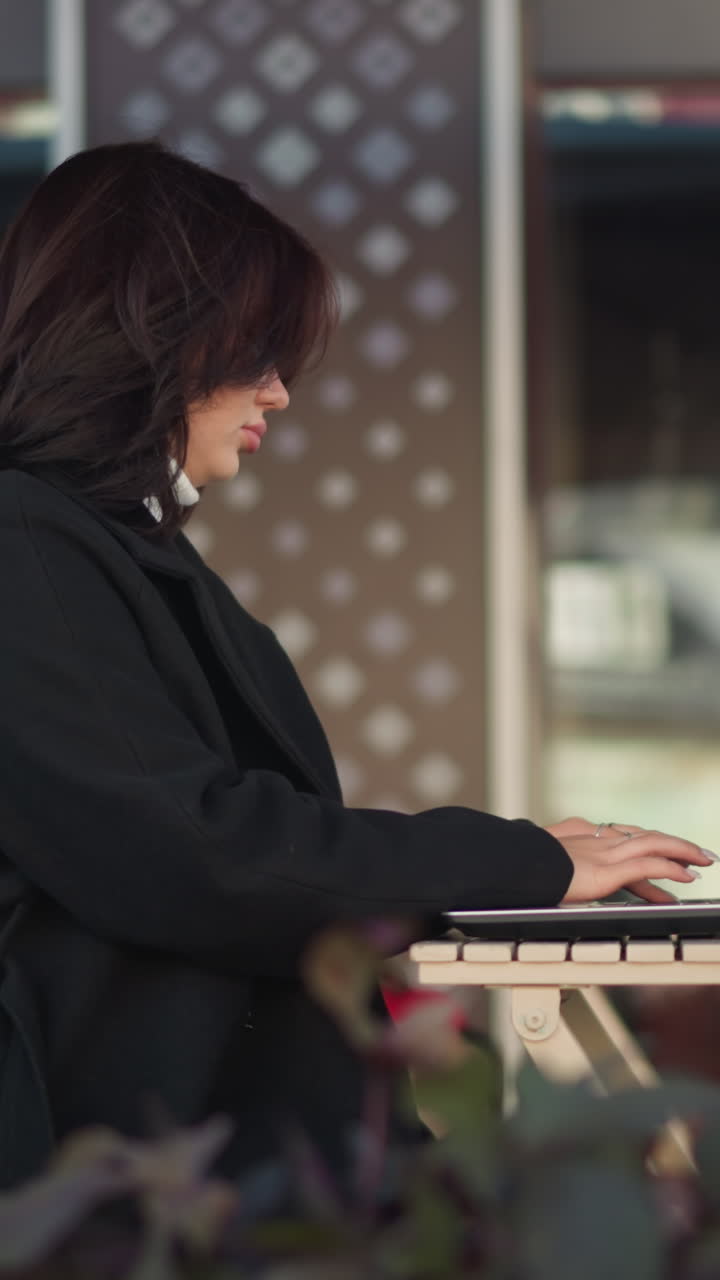 Side view of a professional woman typing on laptop outdoors in urban setting, dressed in a chic black coat, with glass building reflecting nearby structures, decorative flowers close to her