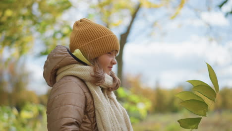 close up of cute girl standing while holding yellow autumn leaf in hand wearing mustard beanie beige quilted jacket and knitted scarf under golden tree canopy on leaf strewn