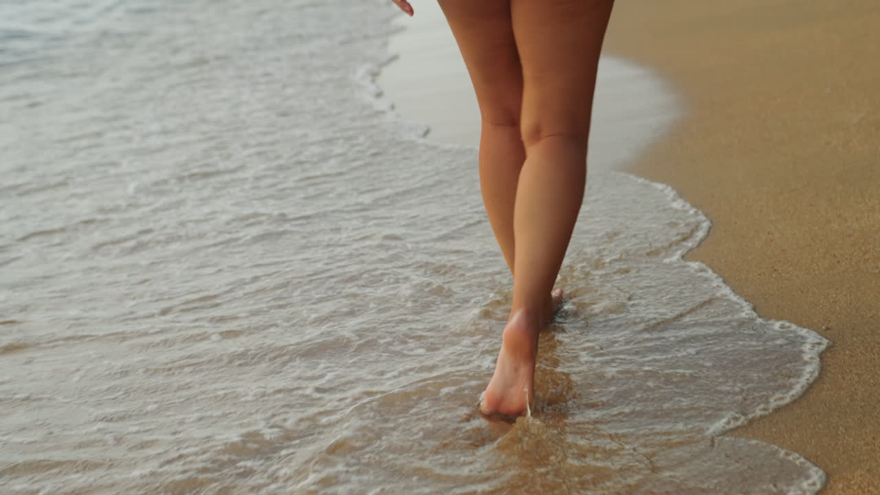 Donna a piedi nudi che cammina sulla spiaggia lungo la sabbia dorata e bagnata con dolci onde che lambiscono, viaggiatrice solitaria che si gode una passeggiata consapevole, impronte che svaniscono nella risacca, luce solare calda e brezza marina che evocano calma.