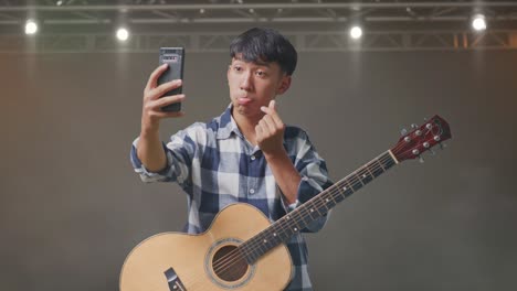 adolescente asiático con selfie de guitarra en su teléfono inteligente mientras está de pie en el escenario