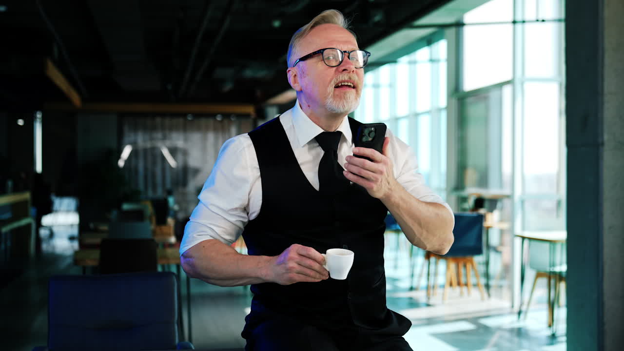 Old man stands in office holding a coffee cup and phone. Businessman scrolls by the smartphone with his nose and tongue.