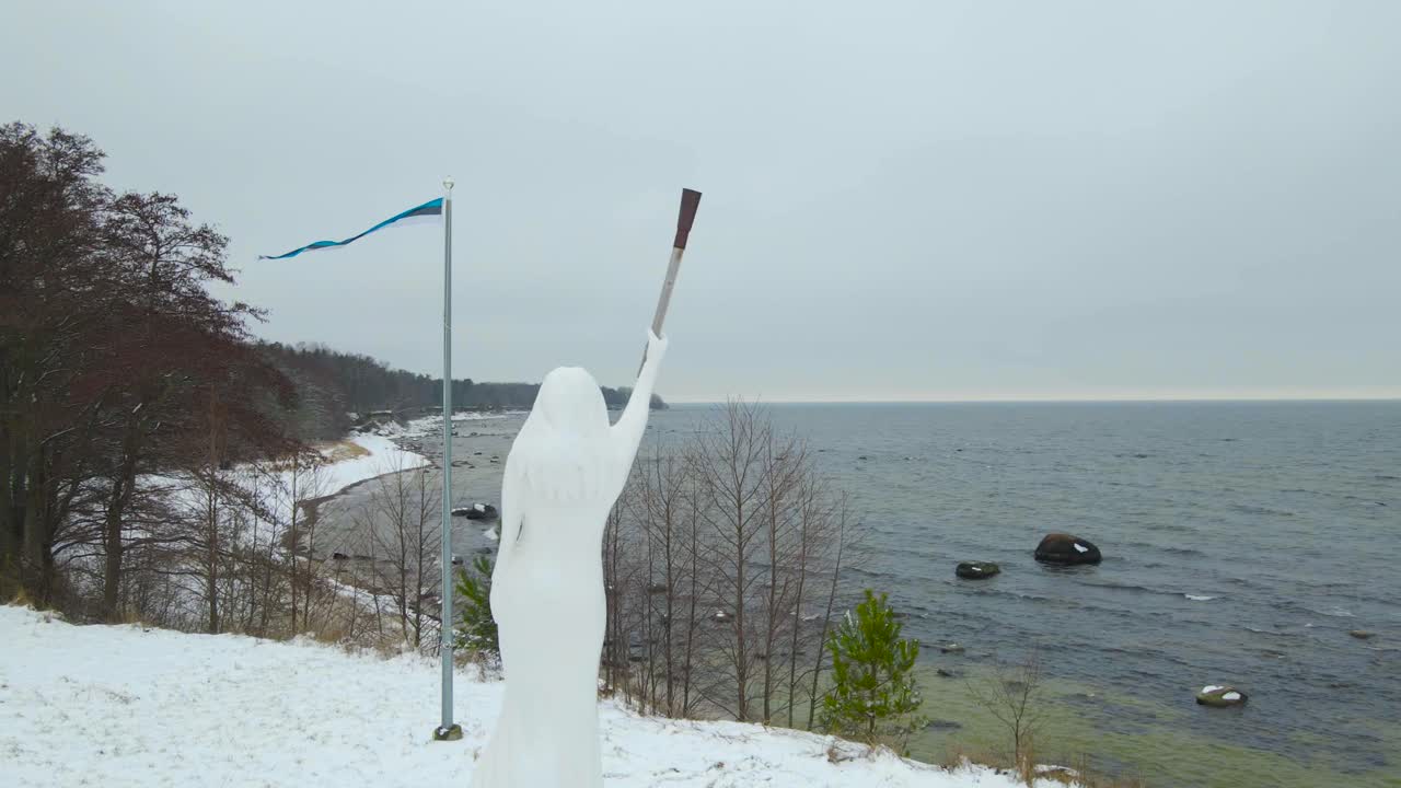 Aerial footage spinning, circling or orbiting around a wooden white Statue of Liberty in Suurupi Estonia during a winter day while a Estonian flag is waving and Baltic sea is visible in the background