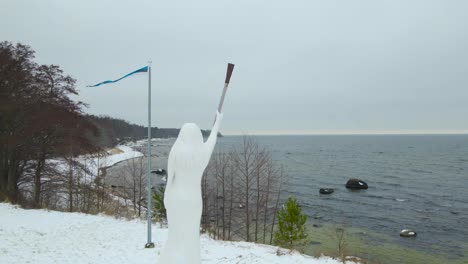 aerial footage spinning, circling or orbiting around a wooden white statue of liberty in suurupi estonia during a winter day while a estonian flag is waving and baltic sea is visible in the background
