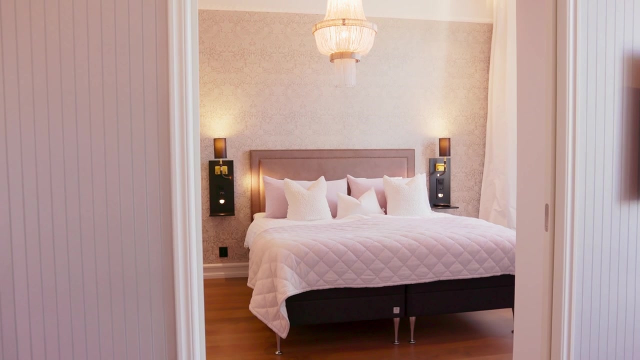 Ampia inquadratura di una camera da letto accogliente con un letto trapuntato al centro, cuscini decorativi, carta da parati a fantasia, applique da comodino, tende trasparenti che incorniciano la luce naturale e una toeletta sulla sinistra.