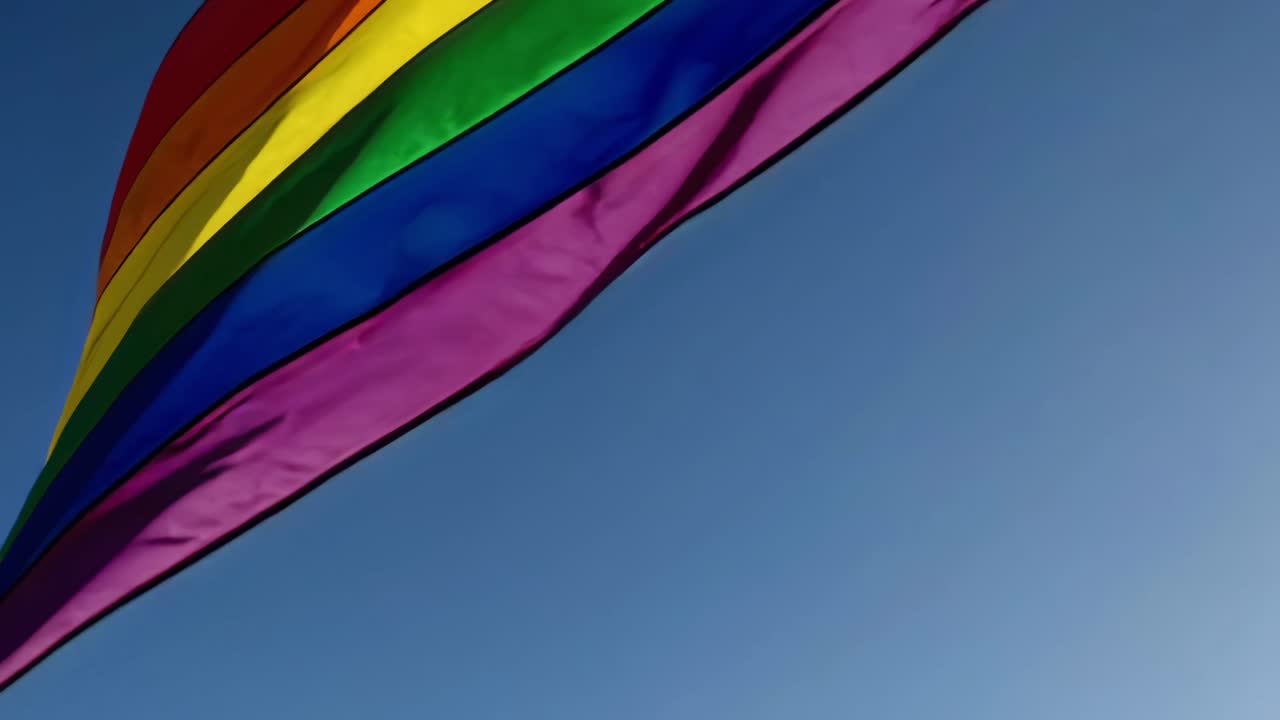 En lav engel fanger et levende regnbueflag, der vajer mod en klar, blå himmel, hvilket symboliserer stolthed