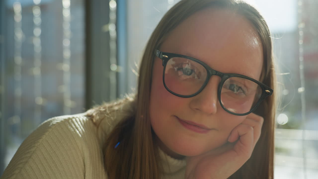 Close up of stylish woman in glasses resting hand on chin while gazing at camera with calm expression. Indoor winter setting with soft light through window decorated with hanging string lights