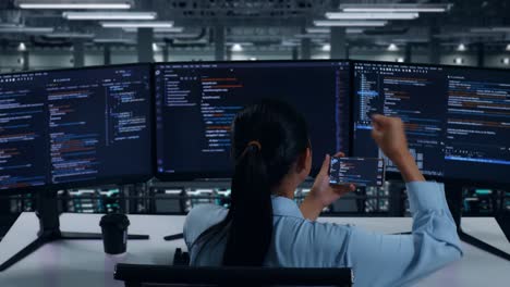 back view of happy asian woman developer looking at code on mobile phone while write code with multiple computer screens in data center