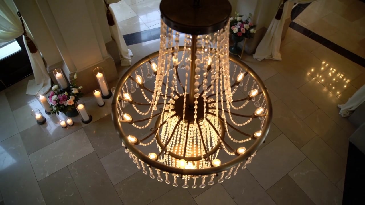Telecamera in movimento che rivela un lampadario di cristallo che riflette la luce sulle piastrelle della sala, mostrando candele e fiori.