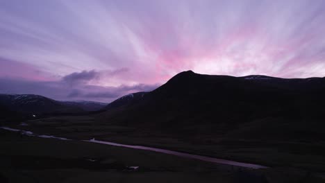 imágenes aéreas de drones de glenshee en escocia durante un intenso cielo rosa y púrpura que cambia de color mirando hacia las montañas silueteadas mientras la puesta de sol se refleja en un río y fuera de las nubes