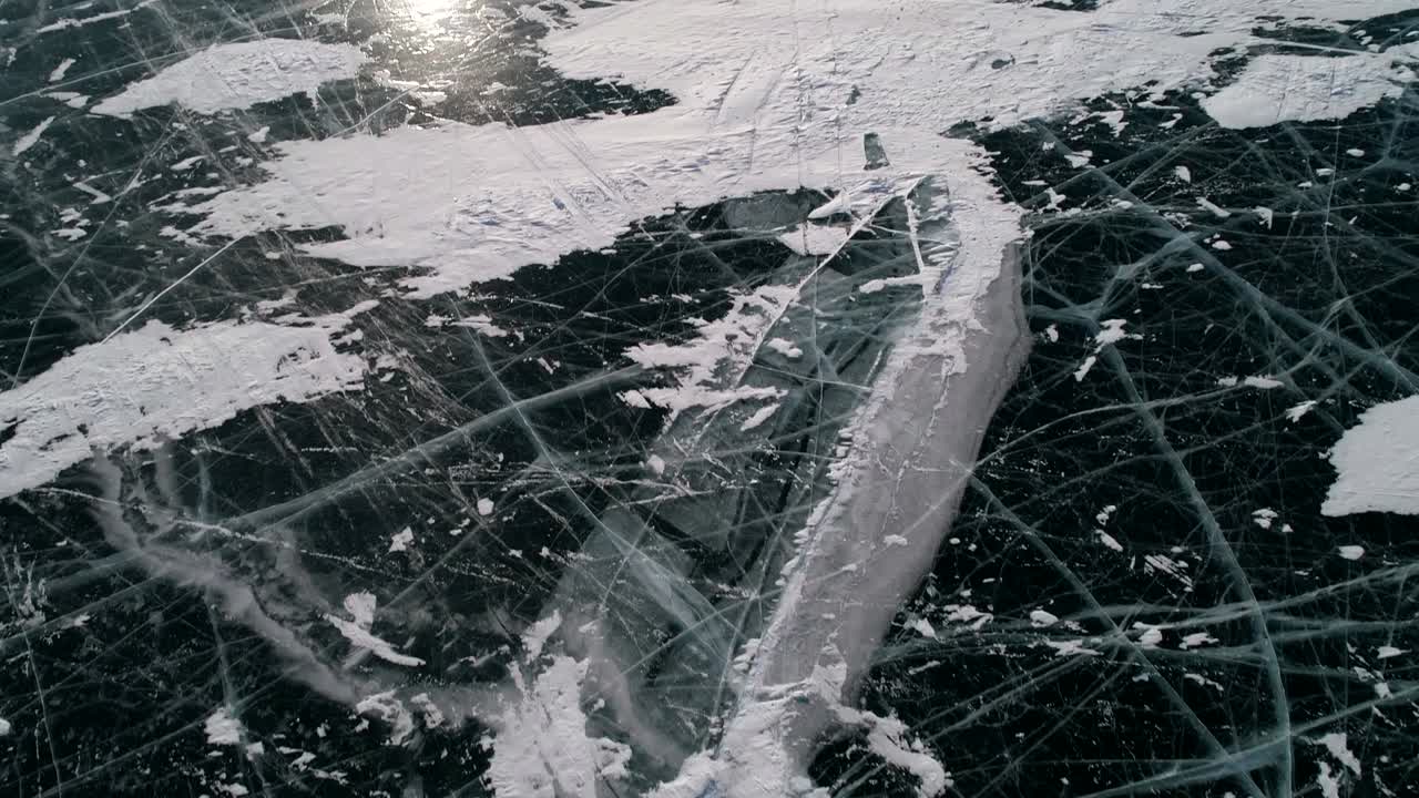 Aerial tilt shot of cracked blue ice and cape Khoboy, Olkhon island. Tall rocks in frozen lake Baikal with many people and cars around. Popular touristic destination. Winter landscape. Panoramic view