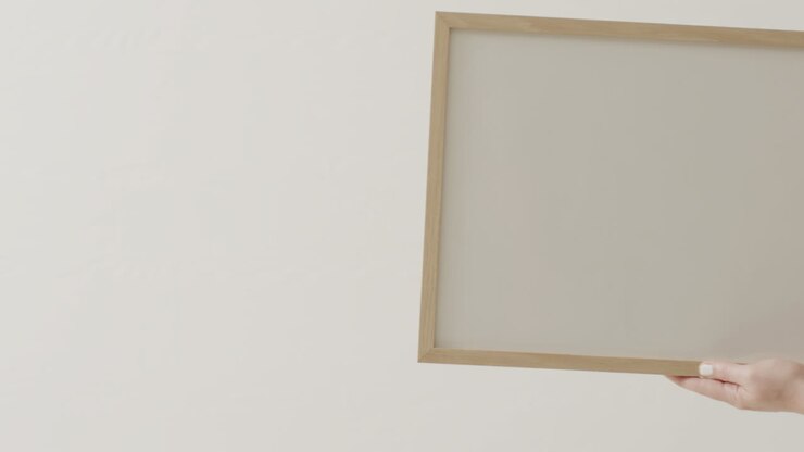 Hand einer hellhäutigen Frau, die einen Rahmen mit Platz für Text auf weißem Hintergrund vor einer weißen Wand hält.