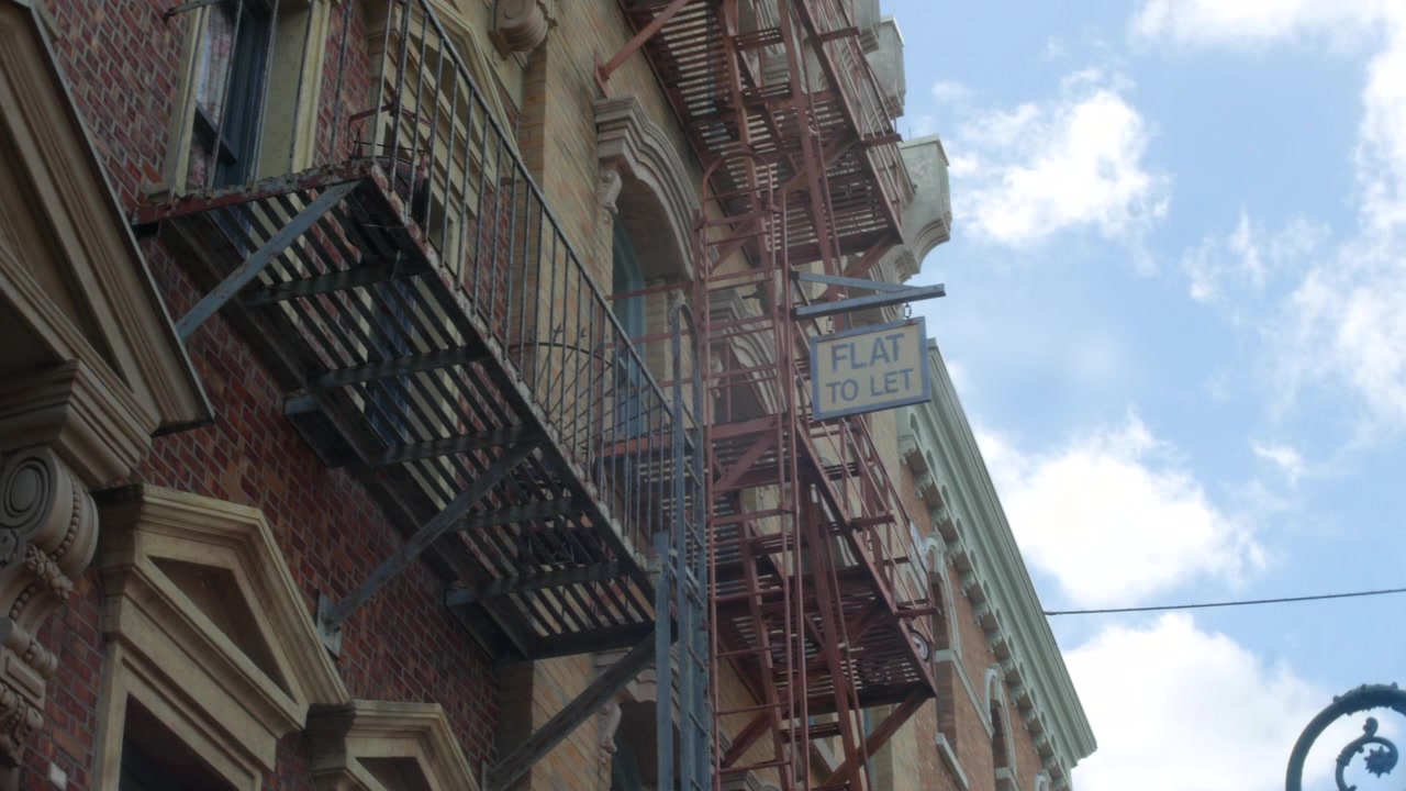Scale antincendio di un edificio che salgono facciate di appartamenti in mattoni accanto a un cartello 'appartamento in affitto'. Vista urbana dal basso che mostra scale metalliche contro un cielo nuvoloso e luminoso