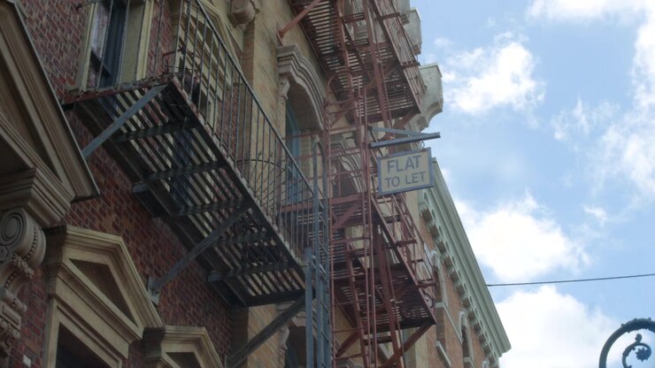 Scale antincendio di un edificio che salgono facciate di appartamenti in mattoni accanto a un cartello 'appartamento in affitto'. Vista urbana dal basso che mostra scale metalliche contro un cielo nuvoloso e luminoso