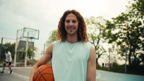 close-up shot of a curly red-haired man in a light-colored t-shirt with a basketball ball posing and looking at the camera outside on a basketball court in summer