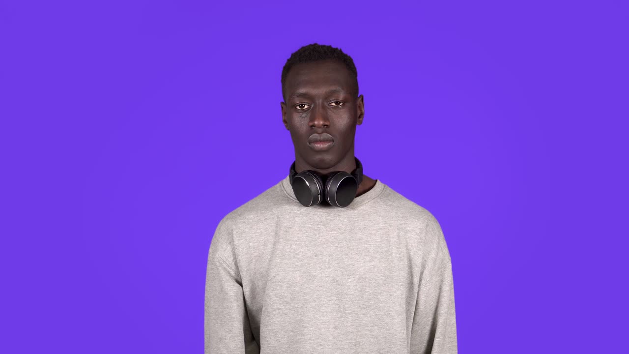 Portrait of young african american man guy in white shirt posing isolated on blue background studio. People emotions lifestyle concept. Showing stop gesture with hand, headphones on neck