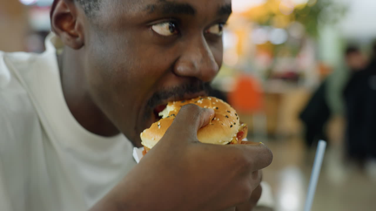 Close up of man hand picking sesame burger from black tray with french fries and drink nearby while seated indoors in food court with relaxed atmosphere and soft lighting in background