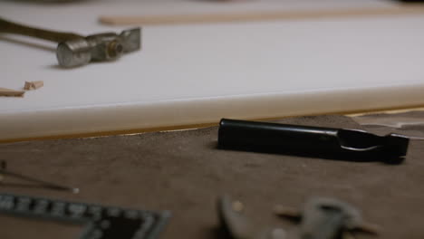 a craftsman's hand reaches for a leatherworking tool on a workbench covered with various tools used for cutting, stamping, and shaping leather.