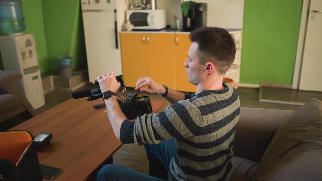 Man adjusting camera on tripod with microphone in home studio, preparing for video production work. Indoor workspace with green background, clock, and festive decorations visible