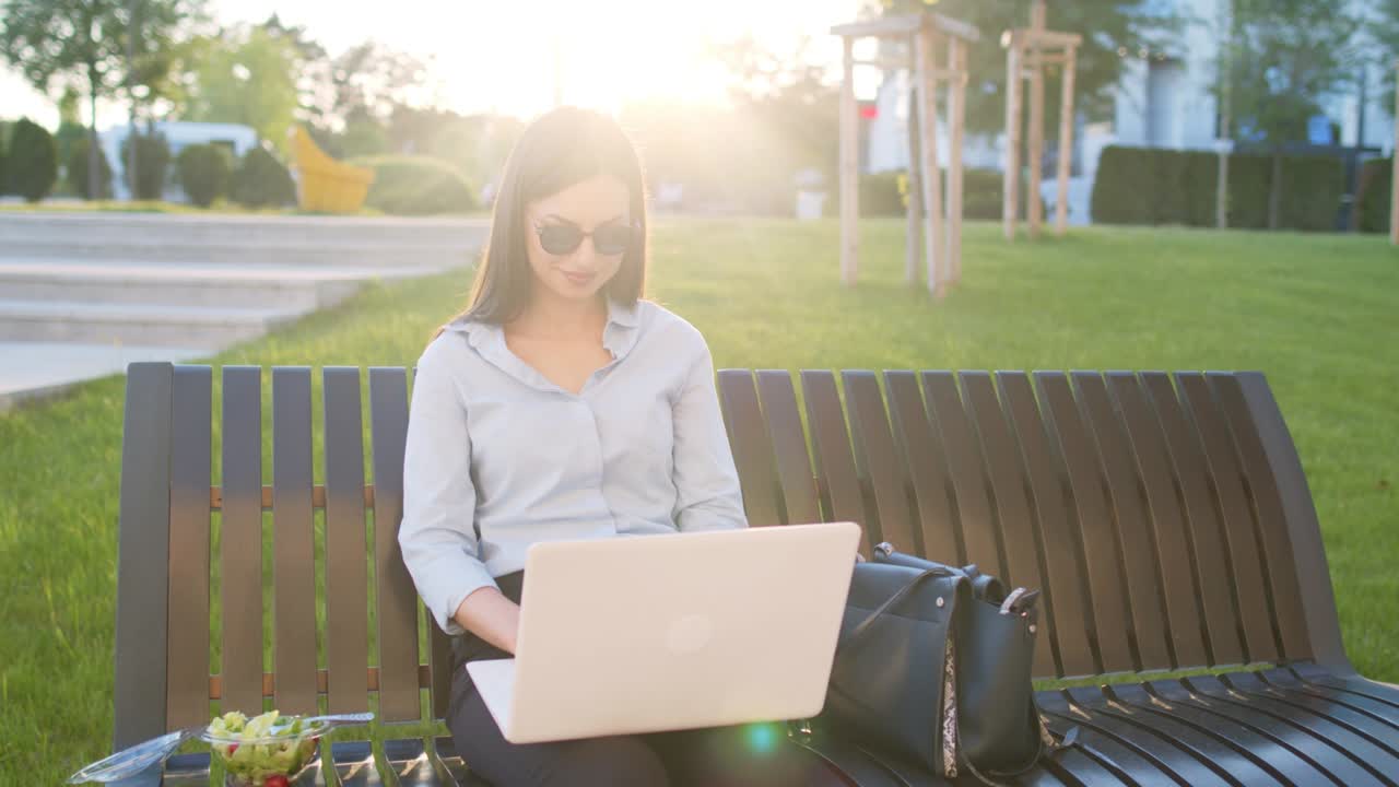 Una donna d'affari è seduta su una panchina all'aperto e usa il laptop per lavorare fuori dall'ufficio e mangia un'insalata.