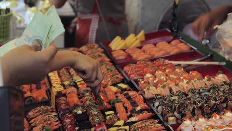 A-steady-close-up-shot-of-people's-hands-while-buying-japanese-sushi-in-a-traditional-thai-street-market,-Thailand