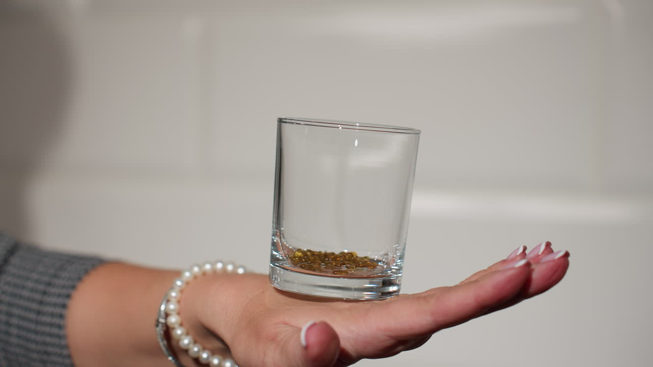 clean glass with beads inside placed on the back of a hand adorned with pearl bracelets, white wall tiles in the background, showcasing delicate hand movement and elegant kitchen decor