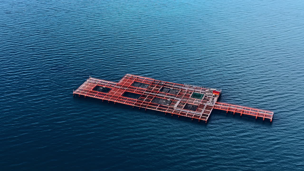 Red rectangular fish farm structure surrounded by ripples. Floating aquaculture platform in open blue sea water. Aerial view