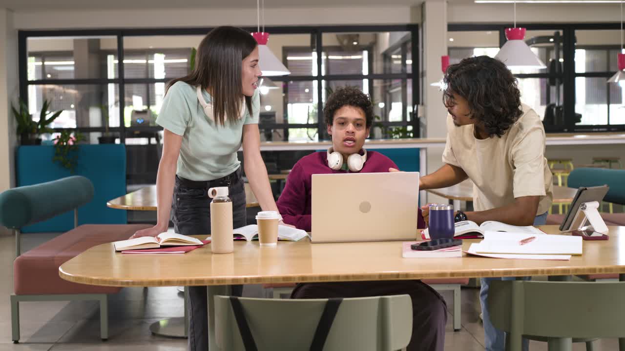Diverse students leaning in at study table after seated student reacting, explaining laptop notes