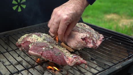 two pieces of lamb meat are being grilled on a coal fired bbq, person is adding wood chips for flavor