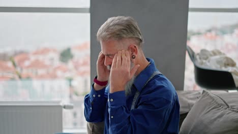 Side-view-of-an-elderly-man-with-gray-hair-and-a-lush-beard-in-a-blue-shirt-kneads-and-massages-his-temples-for-pain-in-his-head-while-sitting-on-the-sofa-in-a-modern-apartment