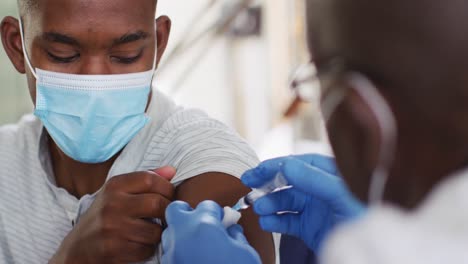 african american senior male doctor giving covid vaccine to male patient in home, wearing face masks