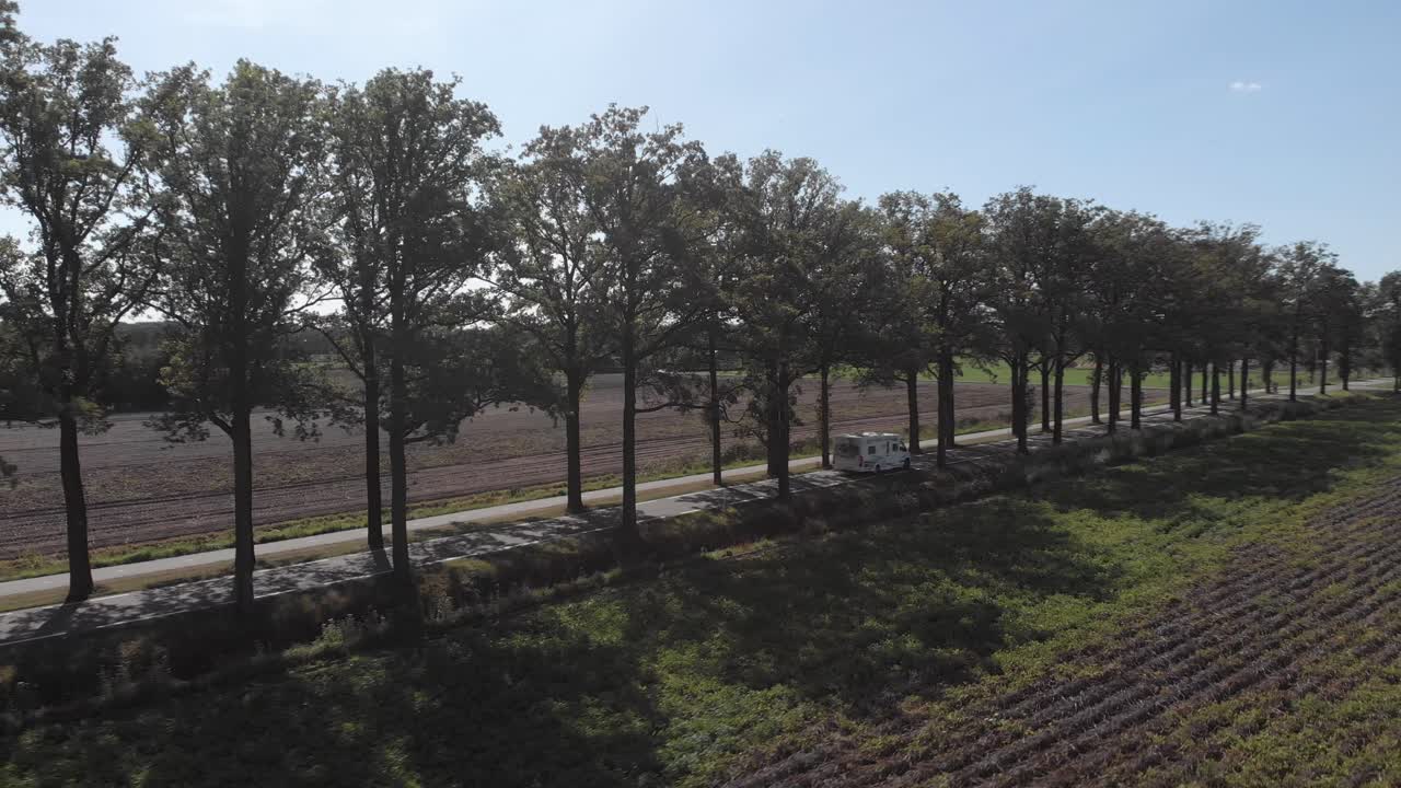 Aerial view of a backlit asphalt country road in The Netherlands with a row of trees backing off and following an approaching camper passing by and disappear into the distance