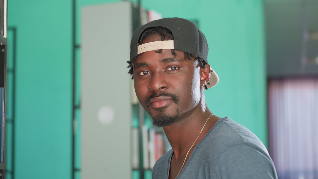 Close up of student wearing cap standing near book shelf in library, turning head toward camera with calm expression, surrounded by rows of books on blurred background