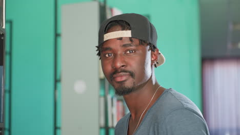 close up of student wearing cap standing near book shelf in library, turning head toward camera with calm expression, surrounded by rows of books on blurred background
