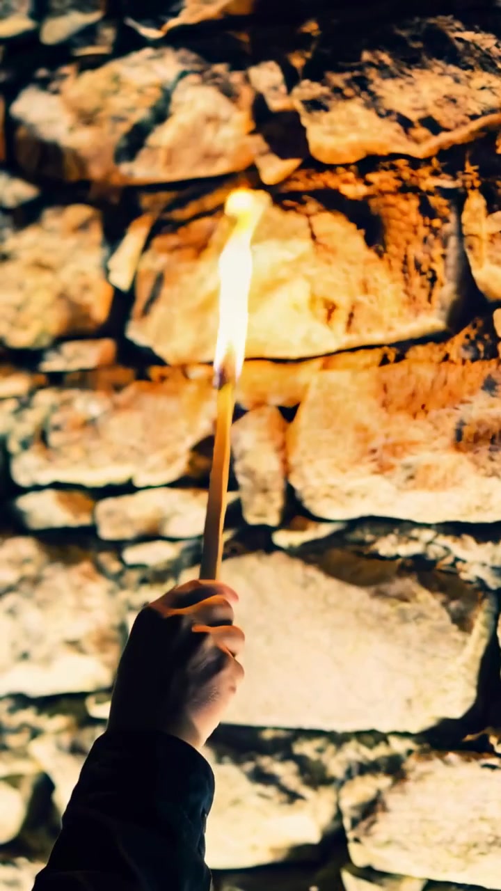 Video verticale: Mano con manica scura che solleva un fiammifero acceso in una cantina di pietra per illuminare la muratura.