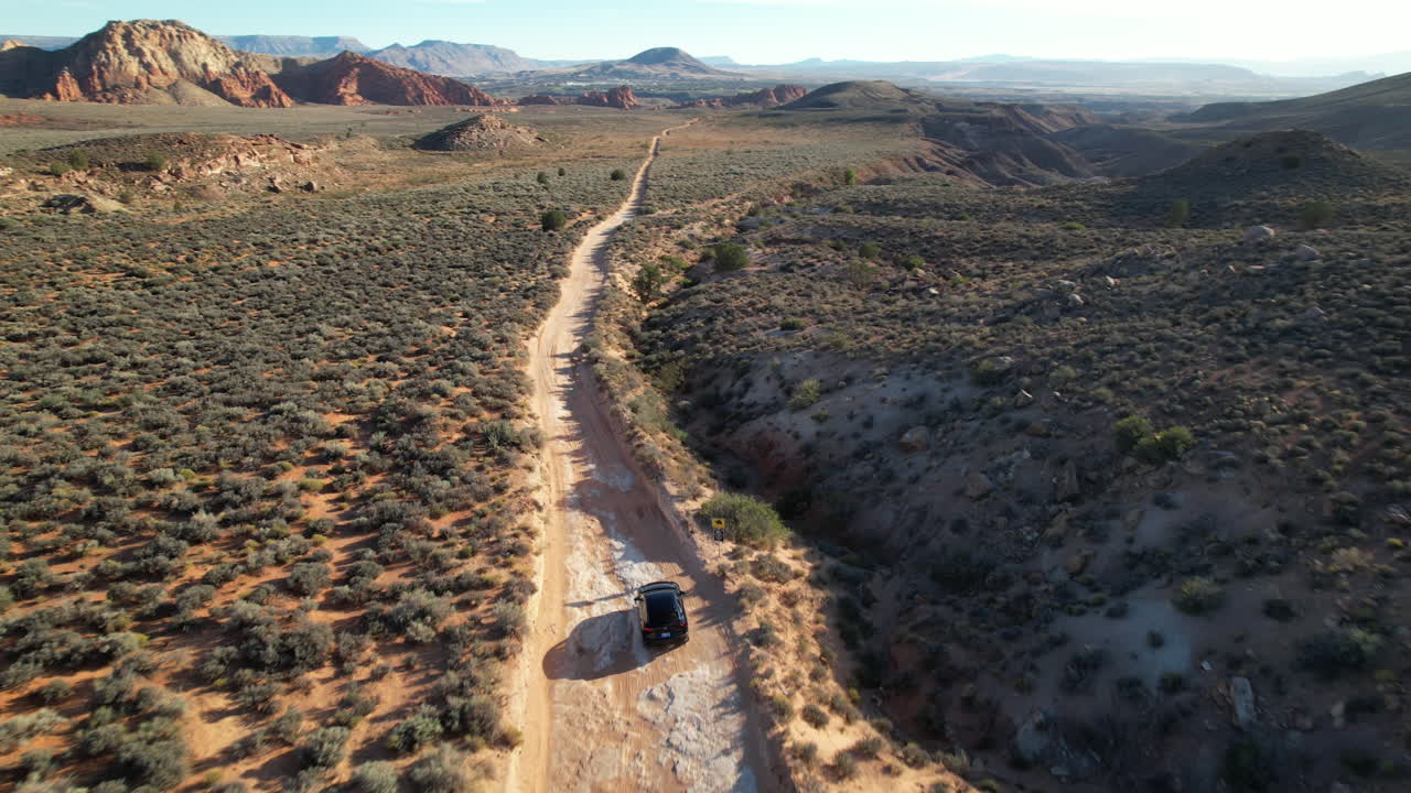 Aerial drone shot tracking an SUV on a rugged dirt road in the Utah desert, surrounded by barren landscapes, shrubs, and distant rocky formations under clear skies.
