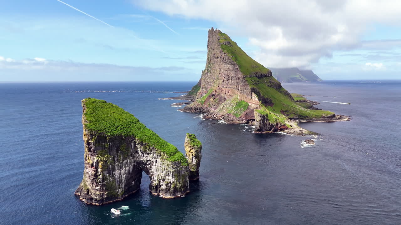 Cinematic aerial view of Drangarnir sea stacks rising dramatically from the Atlantic Ocean near Vágar, Faroe Islands, showcasing rugged cliffs, lush green slopes, and misty Nordic seascape