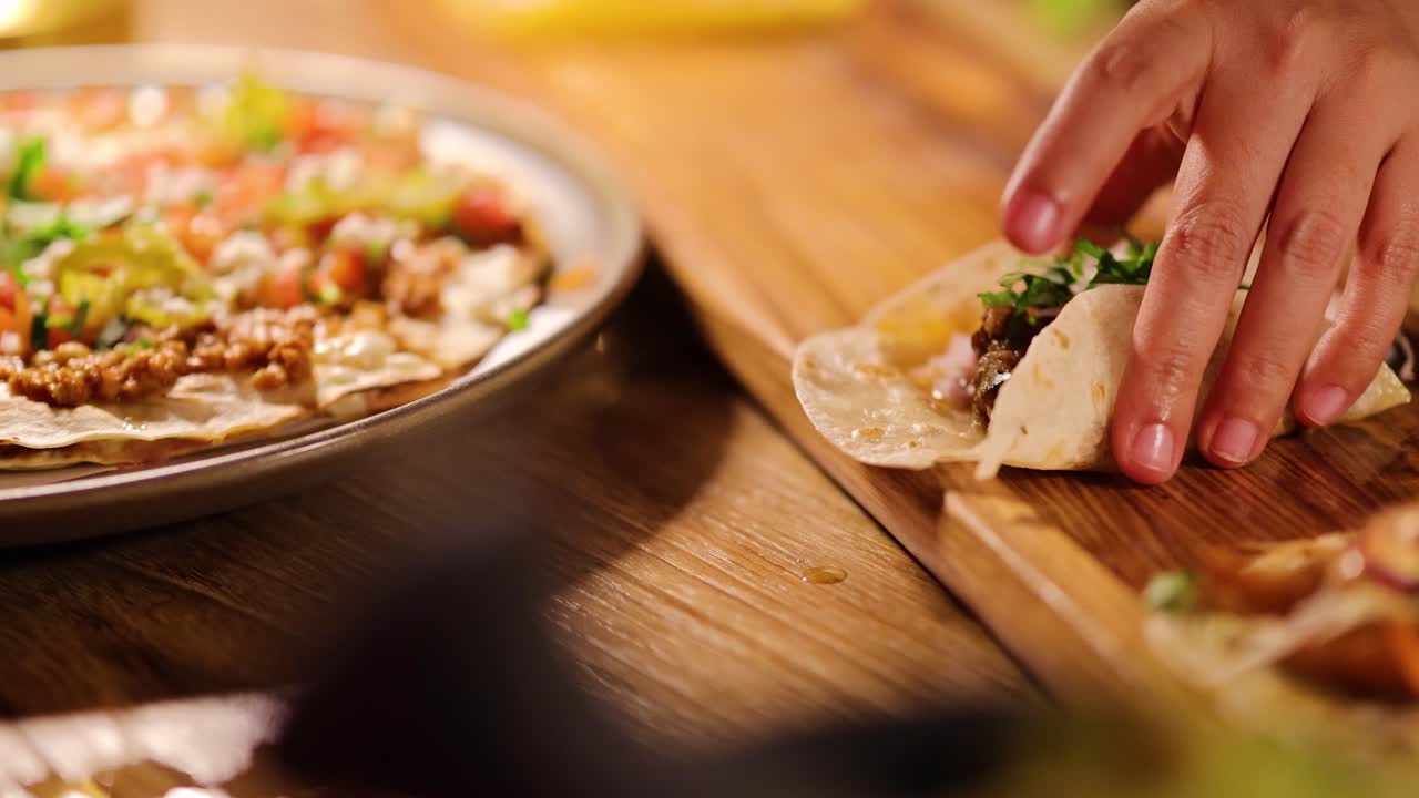 Hand lifts taco from wooden board beside tostada pizza, warm ambient lighting, shallow focus