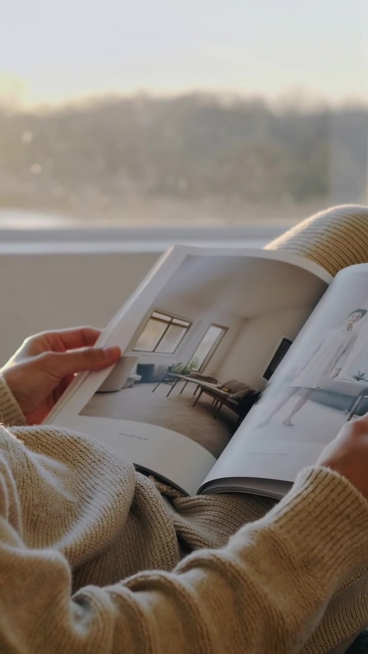 Video verticale: Mani con maglione color crema che sfogliano una rivista di design in grembo a casa, con la finestra retroilluminata.