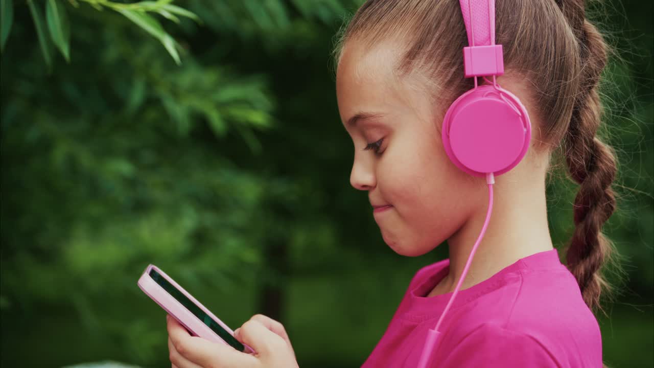 A Young Girl Enjoying Music and Technology Outdoors While Wearing Pink Headphones and Engaged with Her Phone in a Peaceful Natural Setting