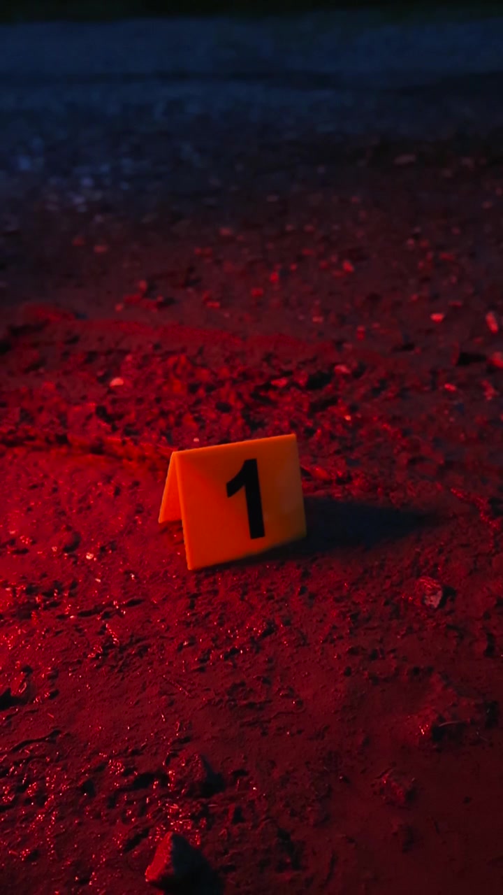 Un segnalatore di prova giallo con il numero uno si trova su terreno fangoso accanto a un'impronta di pneumatico di notte, illuminato dalle luci lampeggianti rosse e blu alternate di un veicolo della polizia.