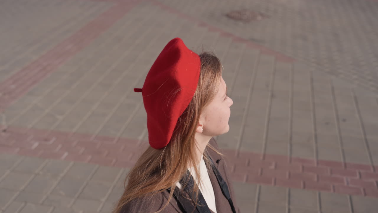 Ung dame med baret smiler. Afslappet portræt af smilende kvinde i gadeomgivelser med blødt lys. Ung kvinde i rød baret smiler blidt med gadefliser som baggrund og afslappet tøj.