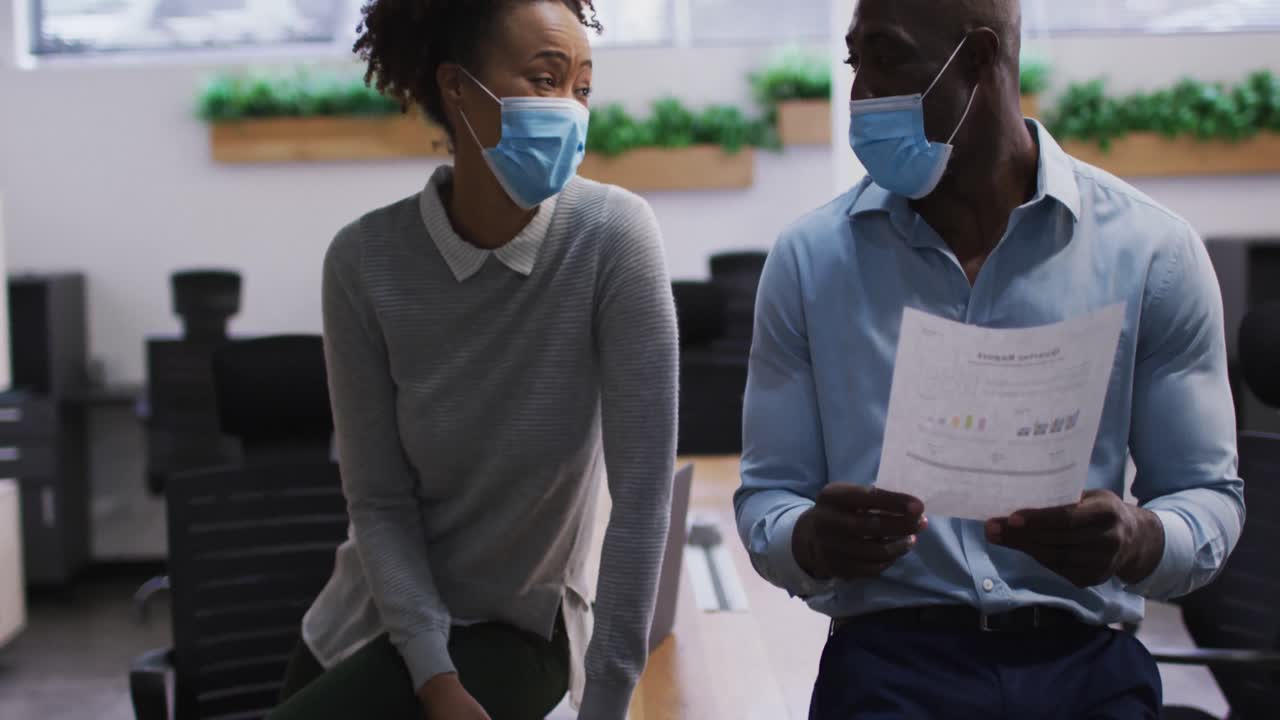 Diverse male and female business colleagues in face masks talking and looking at documents in office