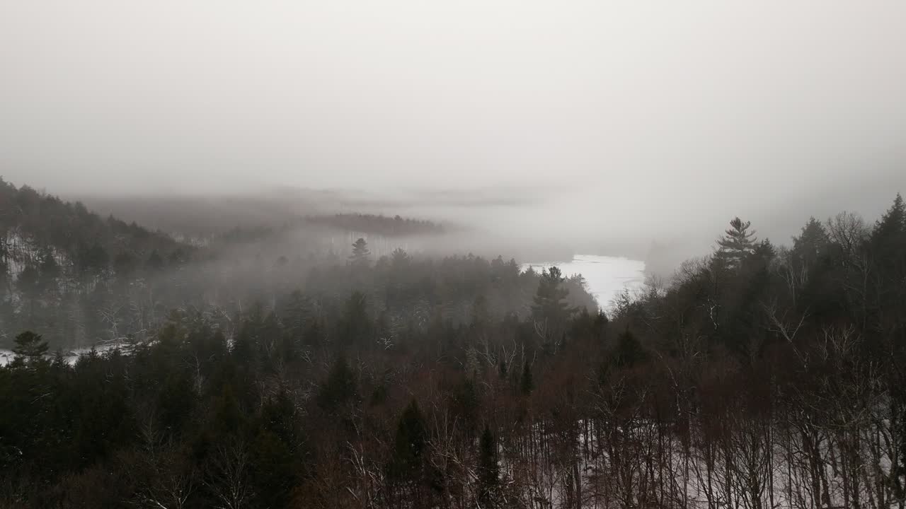 A serene aerial view of a winter forest blanketed in fog, with snow-covered treetops and a distant, partially visible frozen river. The atmosphere exudes peace, mystery, and natural beauty.